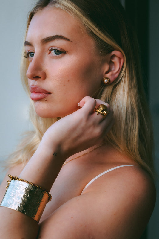 Woman wearing gold jewelry, including earrings and a bracelet, against a neutral background.